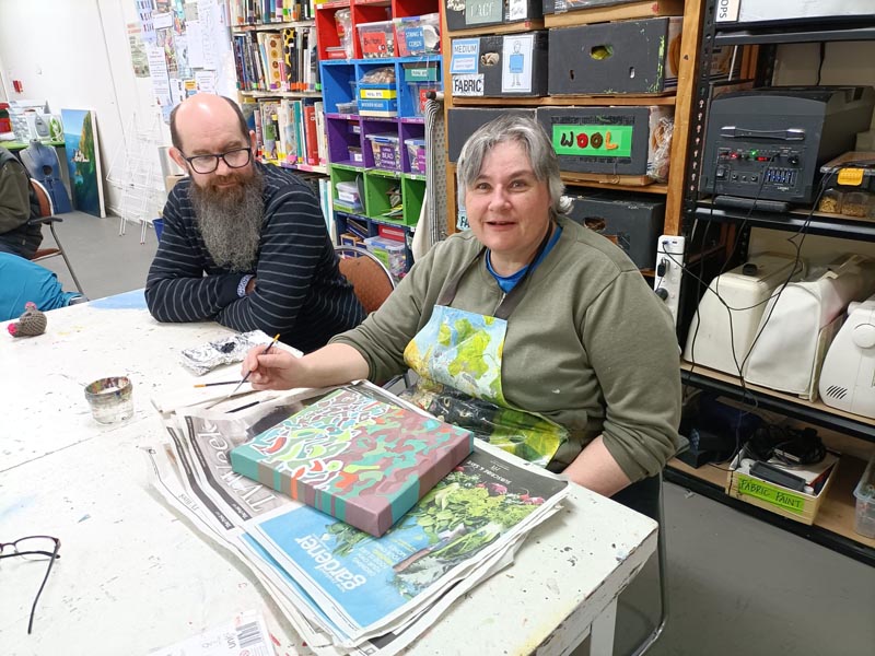 Andrew Mitchell and Emma Gregory seated at a table. Emma has a paint brush in her hand and is working on a painting.